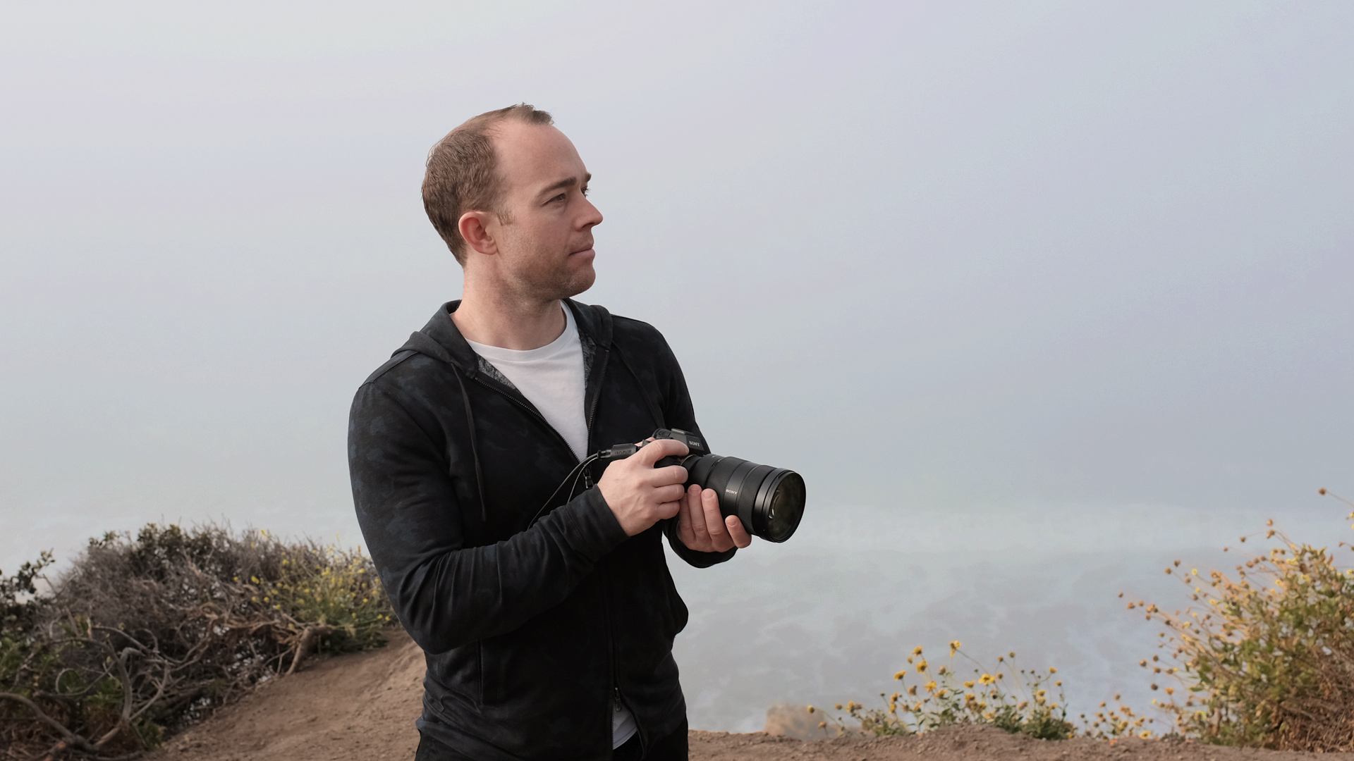 Anthony Furlong on a coastal ridge holding a camera, fog behind him